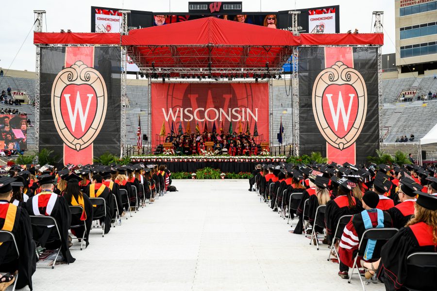 Graduates seated at Camp Randall Stadium facing a commencement stage decorated with banners and flags, and accompanied by speakers and academic faculty and staff.