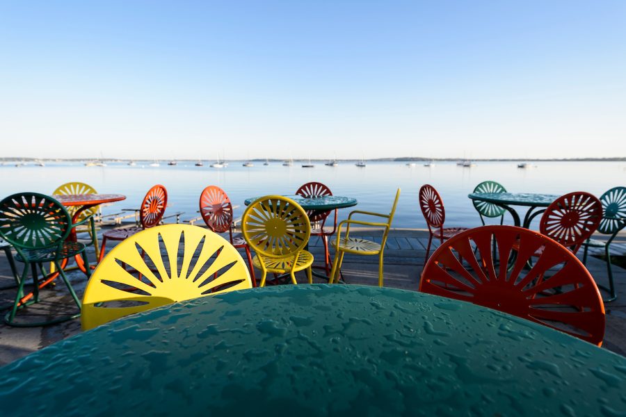 Dew covers colorful sunburst Terrace chairs and tables with lake in background during sunrise.
