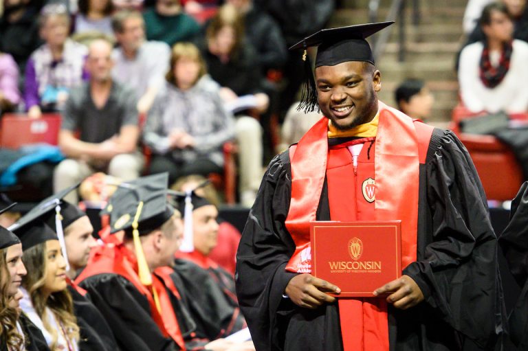 UW graduate in academic attire smiling while holding diploma cover.