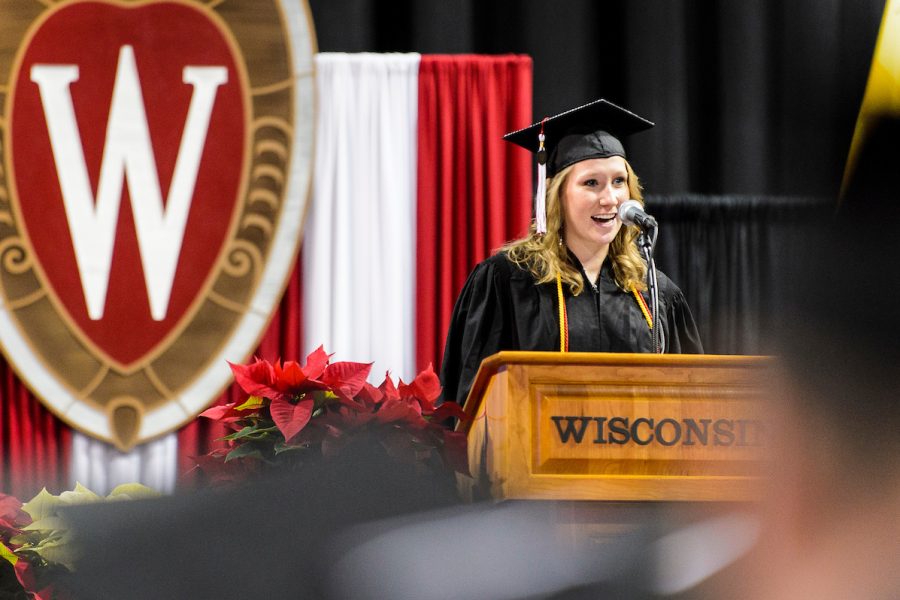 Graduate speaking at podium on stage with UW–Madison crest and banners in background.