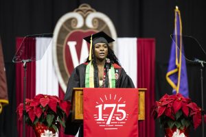 Student dressed in academic attire speaking at podium decorated with UW–Madison 175th logo and university crest in background.