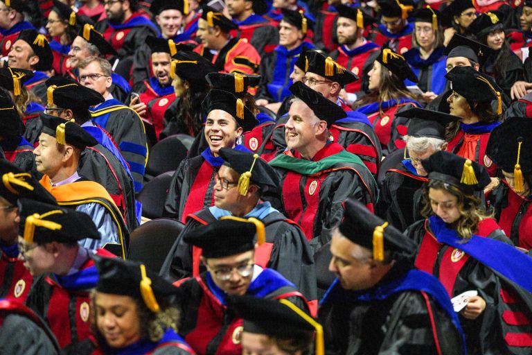 Large group of graduates seated wearing black and red academic attire and blue stoles.