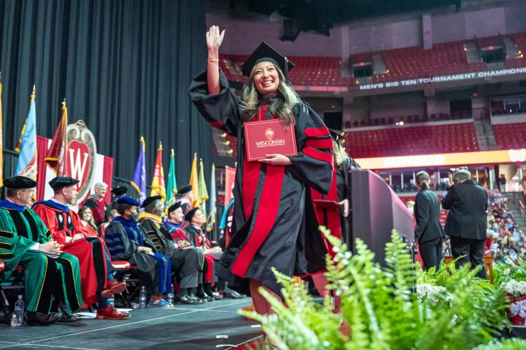 Graduate walking across stage holding red diploma cover smiling and waving.