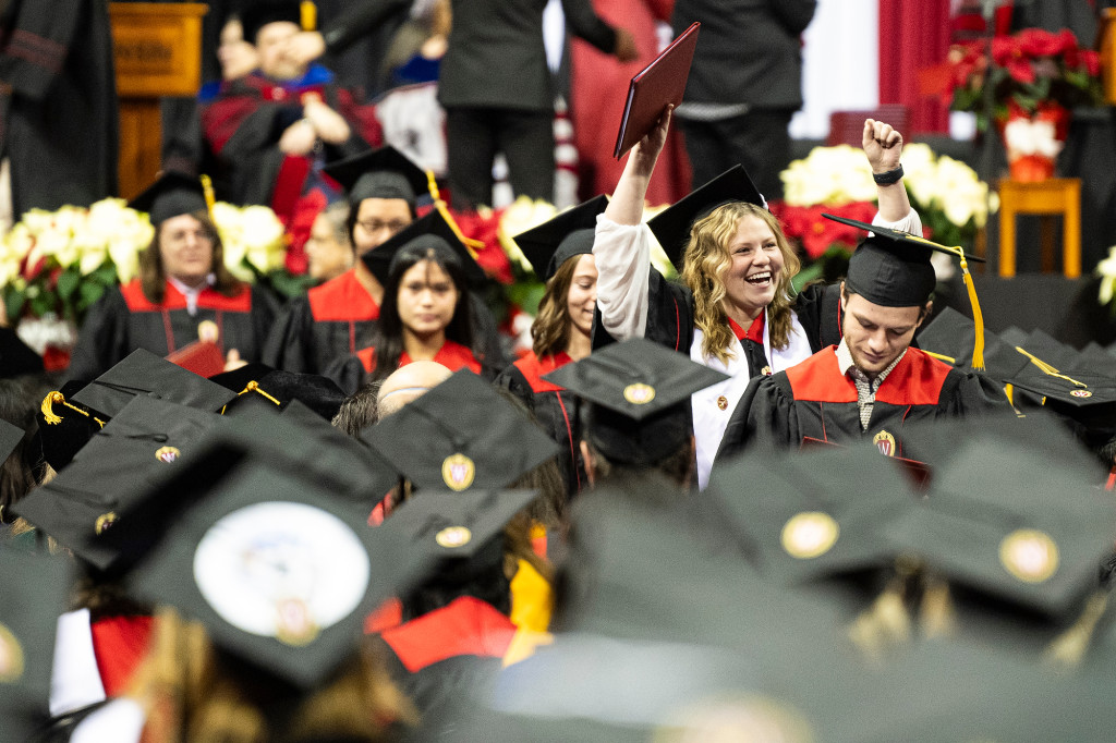 Group of new graduates wearing black and red academic attire smile while holding up diploma covers.