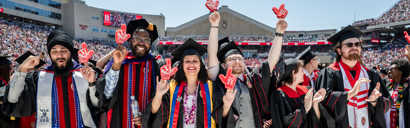 A group of smiling graduates in black caps and gowns with red and white accents wearing red foam fingers celebrate at a graduation ceremony.