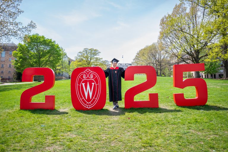 Graduate wearing cap and gown posing outdoors standing in between the numbers 2025.