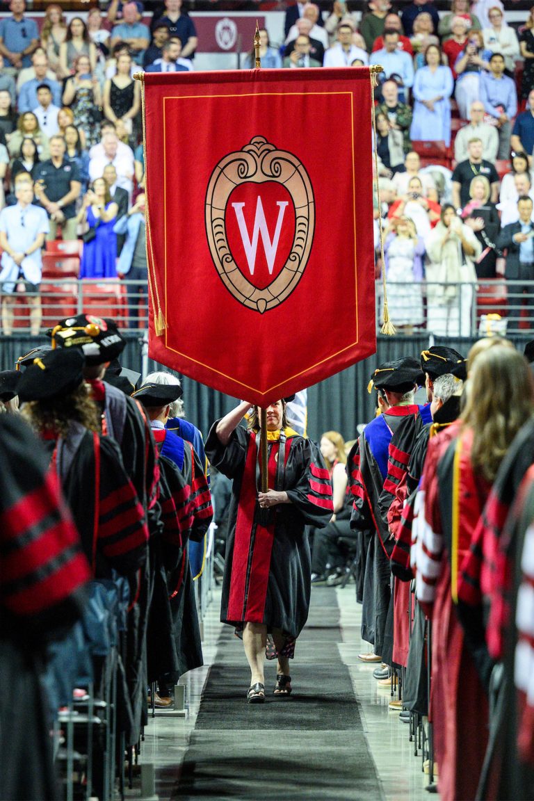 A flag bearer carries a red UW crest banner while graduate candidates stand along both sides with guests in the background.