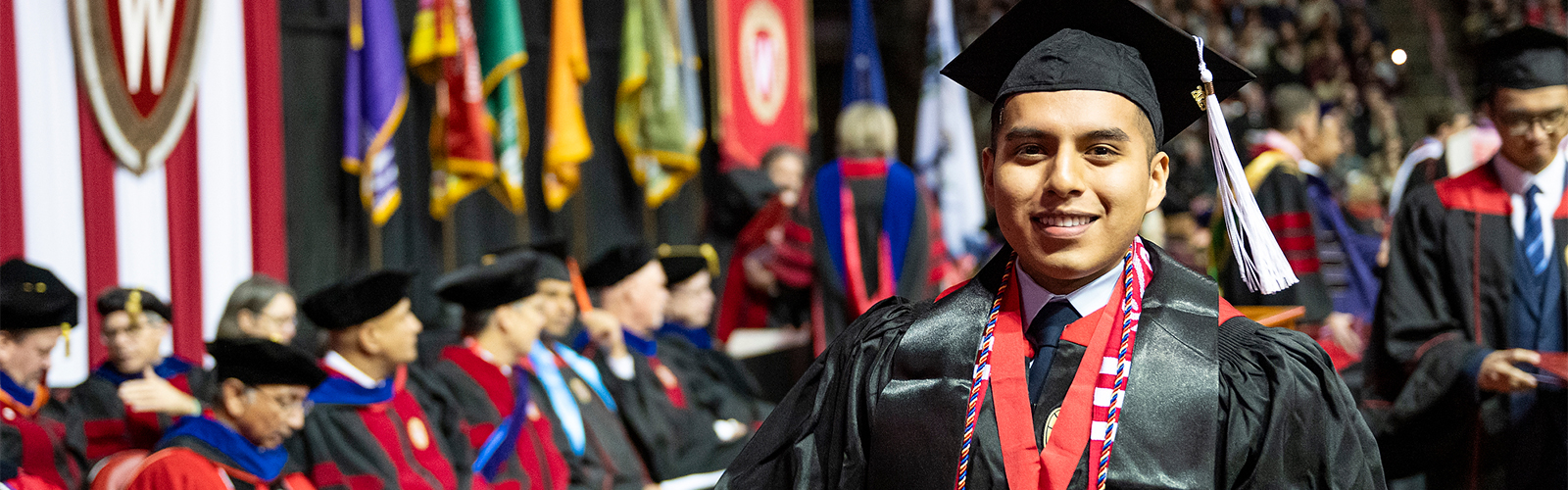 Close-up of a recent graduate in commencement attire walking across the stage, with banners and commencement officials in the background.