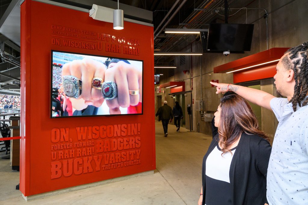 Large display monitor showing two fists wearing championship rings, viewed by two people.
