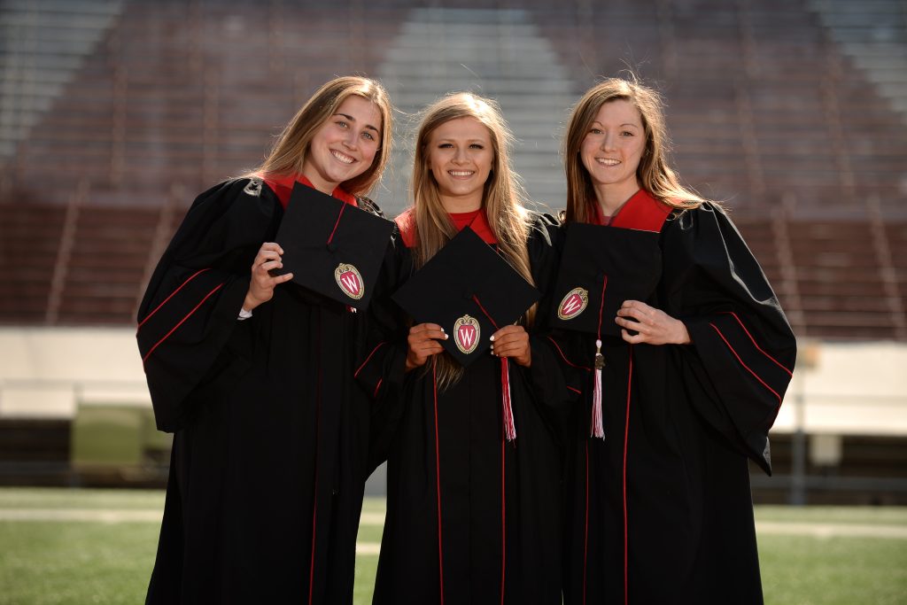 Three UW–Madison students wearing caps and gowns stand on the 50 yard line of Camp Randall. 