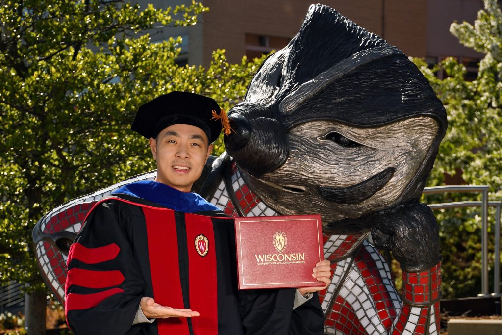 A UW–Madison graduate poses with the Bucky Badger statue wearing cap and gown and holding a diploma cover. 