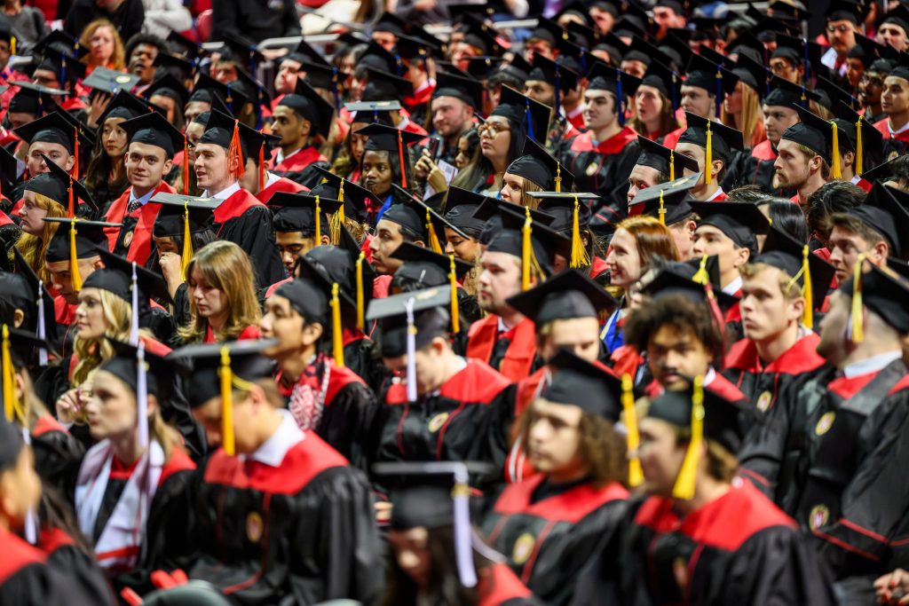 A large group of graduates seated, wearing black caps and gowns with red stoles and tassels of various colors.