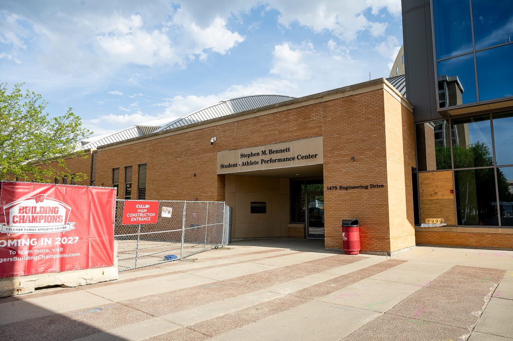 Clouds float in a blue sky above the Student-Athlete Performance Center, adjacent to which is a fence blocking the sidewalk. A red sign on the fence notes that it is a construction entrance.