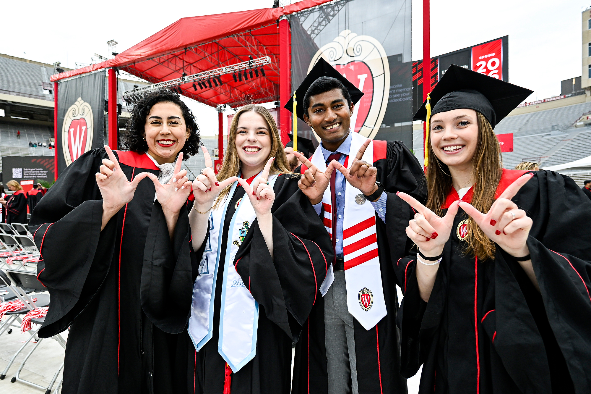 Four graduates in caps and gowns smile and make a “W” with their hands in front of a stadium stage decorated for Spring 2026.